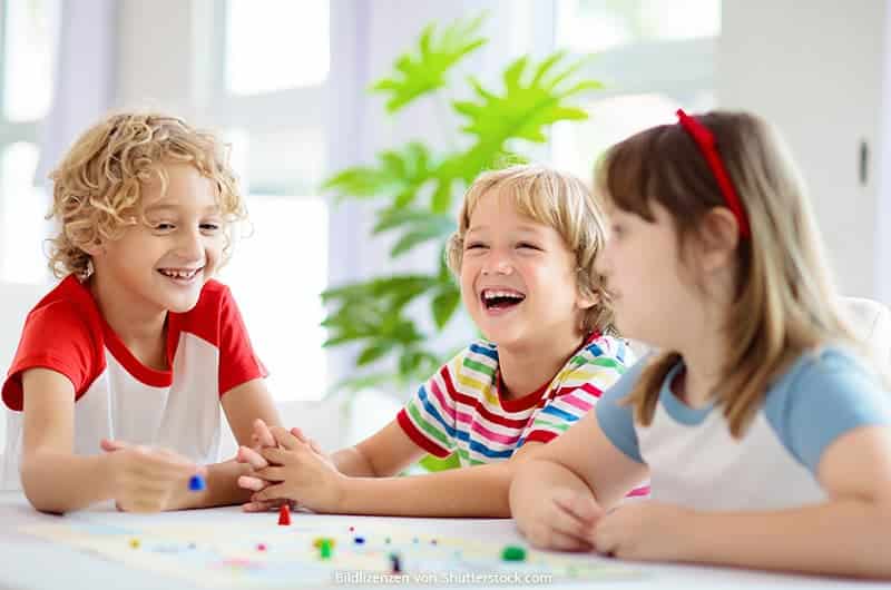 Drei Kinder sitzen lachend am Tisch zuhause und spielen ein Brettspiel