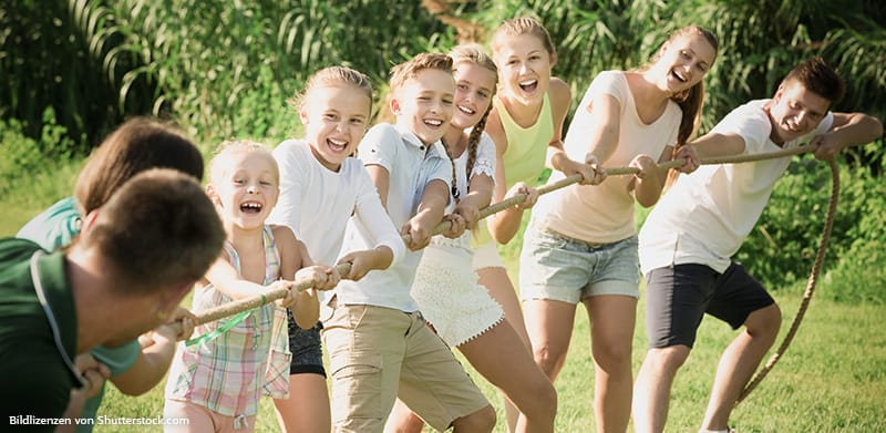 Kinder und Erwachsene spielen Tauziehen im Garten bei Sonnenschein