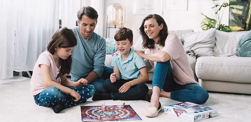 Familie aus Vater, Mutter, Sohn und Tochter spielt gemeinsam im Wohnzimmer vor dem SofaFamilie-aus-Vater-Mutter-Sohn-und-Tochter-spielt-gemeinsam-im-Wohnzimmer-vor-dem-Sofa-das-Brettspiel-Die-Eiskoenigin-Nach-Hause-zusammen-spielen-ASS-Altenburger das Brettspiel Die Eiskoenigin Nach Hause. Zusammen Spielen macht Spaß.