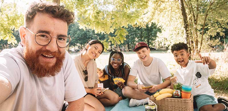 Gruppe von jungen Menschen sitzt im Sommer auf Picknick-Decke im Park, spielt Karten und macht ein Selfie. Sommeranfang, ASS Altenburger