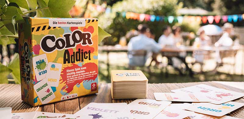 Kartenspiel Color Addict liegt auf Holztisch neben Spielkarten. Im Hintergrund feiert eine Familie im Garten. Sommeranfang, ASS Altenburger
