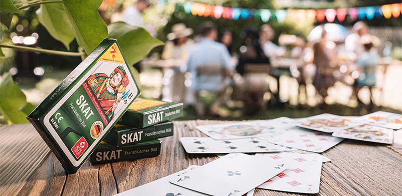 Skatspiel mit französischem Blatt liegt auf Holztisch neben Spielkarten im Hintergrund feiert eine Familie im Garten. Skatstadt Altenburg, ASS Altenburger