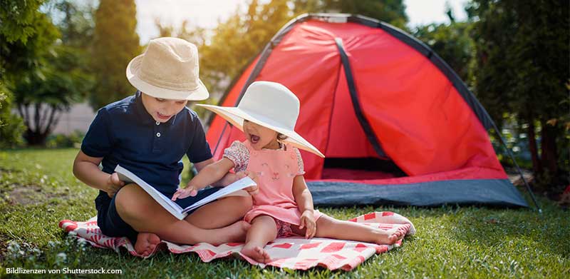 Kinder sitzen auf Wiese vor rotem Zelt und lesen ein Buch. Aktivitäten mit Kindern, ASS Altenburger