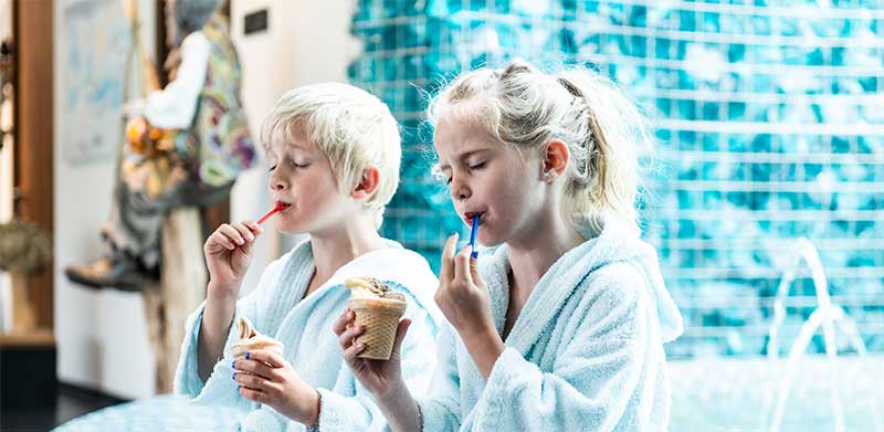 Ein Junge und ein Mädchen sitzen in weißen Bademänteln und mit geschlossenen Augen nebeneinander und essen Softeis.  Familux Resorts, Wellness mit Kindern, ASS Altenburger