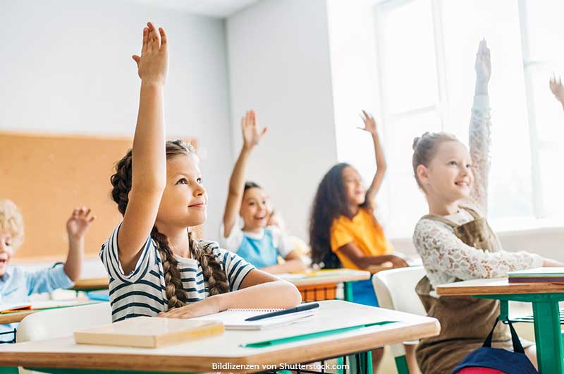 Kinder sitzen an Holztisch im Unterricht, lachen und melden sich. Ehrgeiz, ASS Altenburger