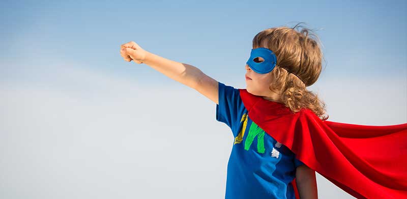 Kleiner Junge steht vor blauem Himmel und trägt eine blaue Superhelden-Maske und einen roten Superhelden-Umhand und streckt die Faust nach oben. Ehrgeiz, ASS Altenburger