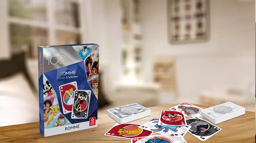 Es ist das ASS Altenburger Disney 100 Rommé Kartenspiel mit Schachtel und Karten vor einem bunten Hintergrund zu sehen.