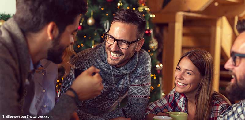 Gruppe von Freunden sitzt im Wohnzimmer vor Weihnachtsbaum und lacht. Weihnachtsmarkt zuhause, ASS Altenburger