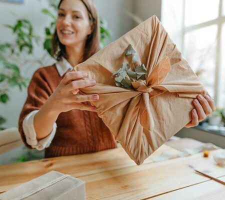 Junge Frau hält Geschenk in die Kamera, das in Stoff eingepackt und mit einem Zweig verziert ist. Nachhaltige Geschenke, ASS Altenburger