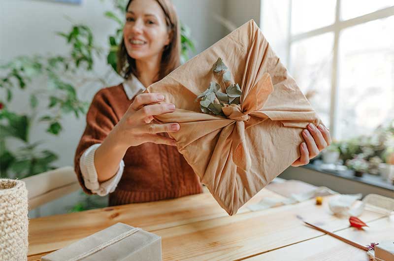 Junge Frau hält Geschenk in die Kamera, das in Stoff eingepackt und mit einem Zweig verziert ist. Nachhaltige Geschenke, ASS Altenburger