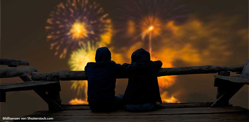 Zwei Personen sitzen auf verschneitem Steg und schauen sich Feuerwerk an. Jahresende, ASS Altenburger