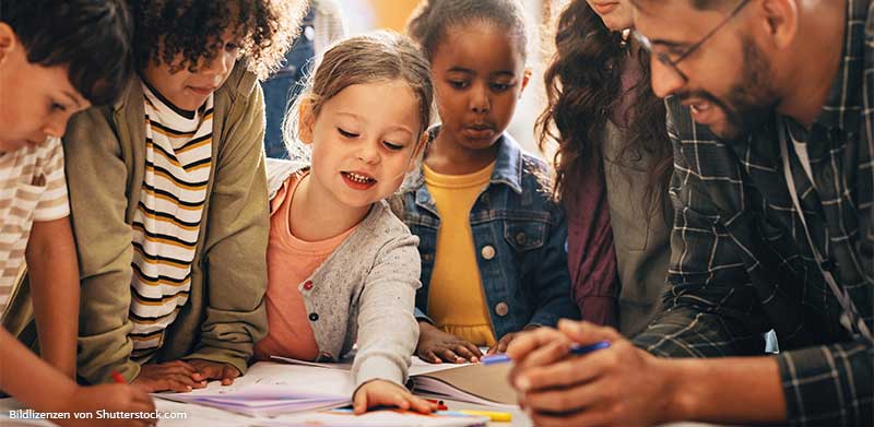 Maedchen-steht-zwischen-fuenf-weiteren-Menschen-am-Tisch-und-zeigt-auf-Papier-in-der-Mitte-Soziale-Kompetenz-bei-Kindern-ASS-Altenburger