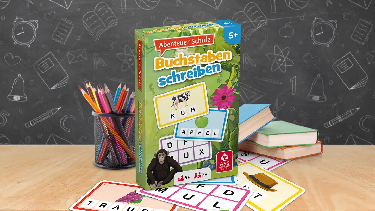 Das Abenteuer Schule Buchstaben schreiben Spiel steht auf einem Schultisch. Unter dem Tisch liegen einige der dazugehörigen Spielkarten. Neben dem Spiel stehen Stifte und ein Bücherstapel. Im Hintergrund ist unscharf eine Tafel zu erkennen.