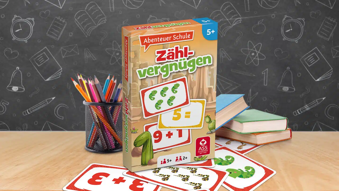 Das Abenteuer Schule Zählvergnügen Spiel steht auf einem Schultisch. Unter dem Tisch liegen einige der dazugehörigen Spielkarten. Neben dem Spiel stehen Stifte und ein Bücherstapel. Im Hintergrund ist unscharf eine Tafel zu erkennen.