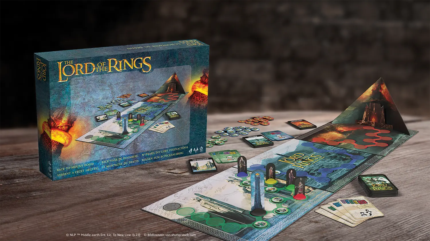 Es sind die Spielkomponenten des Lord of the Rings Mount Doom Spiels auf einem Holztisch zu sehen, im Hintergrund steht die Schachtel.