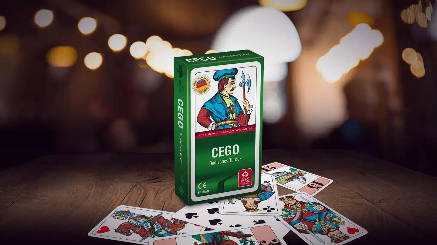Das Cego Kartenspiel steht auf einem Untergrund aus Holz, unter dem Spiel liegen einige der dazugehörigen Spielkarten. Im Hintergrund sind unscharf einige Lichter wie in einer Kneipenbeleuchtung zu erkennen.