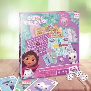 Die Gabby´s Dollhouse Spielesammlung steht vor einem hellem, freundlichem, grünem Hintergrund. Unter dem Spiel sind einige der dazugehörigen Spielkarten zu sehen, neben dem Spiel liegen drei Würfel.