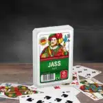 Das Kartenspiel Jass steht auf einem Untergrund aus Holz, unter dem Spiel liegen einige der dazugehörigen Spielkarten. Der Hintergrund ist dunkel.