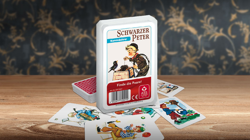 Das Schwarzer Peter Kaminkehrer Kartenspiel steht auf einem Holztisch. Unter dem Spiel liegen einige der dazugehörigen Spielkarten. Im Hintergrund erkennt man unscharf eine mit goldenen Ornamenten verzierte dunkelblaue Wand.