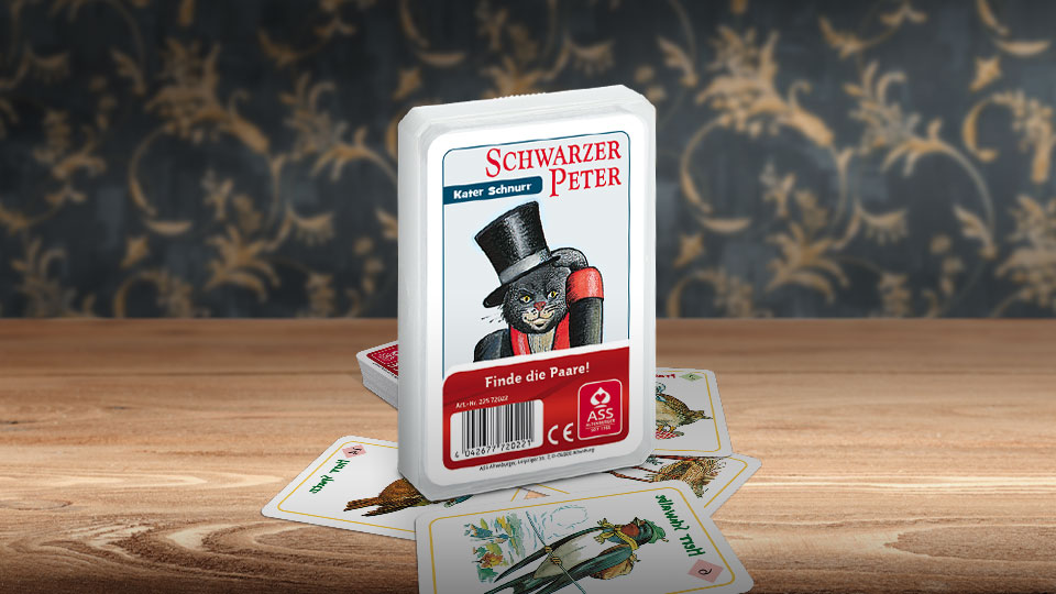 Das Schwarzer Peter Kater Schurr Kartenspiel steht auf einem Holztisch. Unter dem Spiel liegen einige der dazugehörigen Spielkarten. Im Hintergrund erkennt man unscharf eine mit goldenen Ornamenten verzierte dunkelblaue Wand.