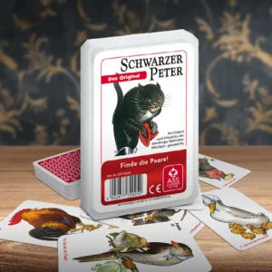 Das Schwarzer Peter Original Kartenspiel steht auf einem Holztisch. Unter dem Spiel liegen einige der dazugehörigen Spielkarten. Im Hintergrund erkennt man unscharf eine mit goldenen Ornamenten verzierte dunkelblaue Wand.