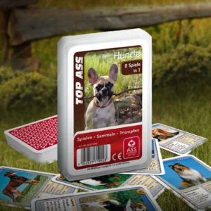 Das Top ASS Hunde Trumpfspiel steht auf einer Wiese, unter dem Spiel liegen einige der dazugehörigen Spielkarten. Im Hintergrund steht ein Zaun der eine anschließende Weide abgrenzt.