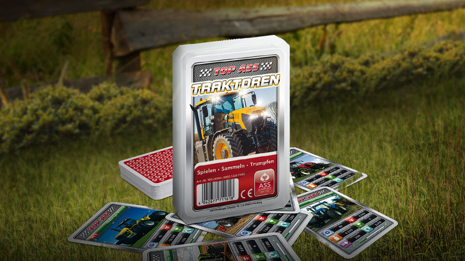 Das Top ASS Traktoren Trumpfspiel steht auf einer Wiese, unter dem Spiel liegen einige der dazugehörigen Spielkarten. Im Hintergrund steht ein Zaun der eine anschließende Weide abgrenzt.