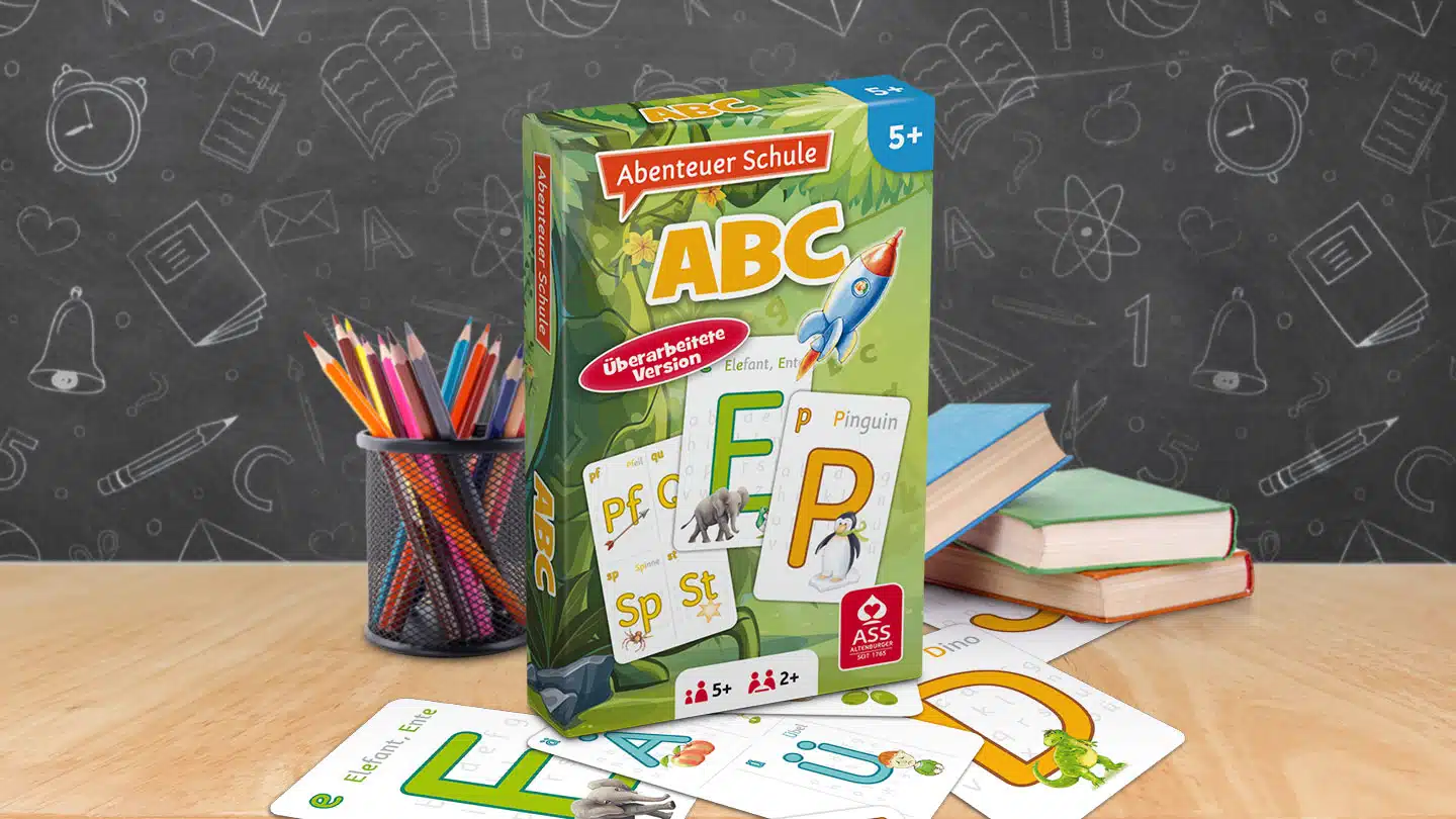 Das Abenteuer Schule ABC Spiel steht auf einem Schultisch. Unter dem Tisch liegen einige der dazugehörigen Spielkarten. Neben dem Spiel stehen Stifte und ein Bücherstapel. Im Hintergrund ist unscharf eine Tafel zu erkennen.