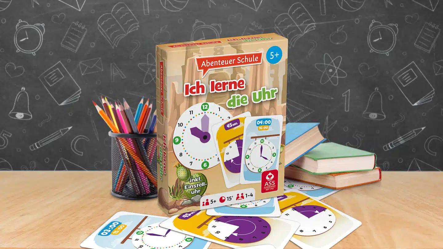 Das Abenteuer Schule Ich lerne die Uhr Spiel steht auf einem Schultisch. Unter dem Tisch liegen einige der dazugehörigen Spielkarten. Neben dem Spiel stehen Stifte und ein Bücherstapel. Im Hintergrund ist unscharf eine Tafel zu erkennen.