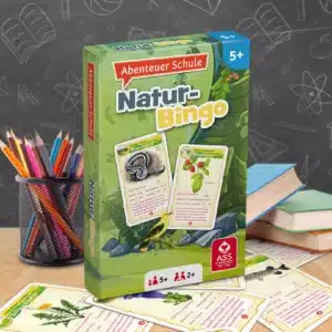 Das Abenteuer Schule Naturbingo Spiel steht auf einem Schultisch. Unter dem Tisch liegen einige der dazugehörigen Spielkarten. Neben dem Spiel stehen Stifte und ein Bücherstapel. Im Hintergrund ist unscharf eine Tafel zu erkennen.