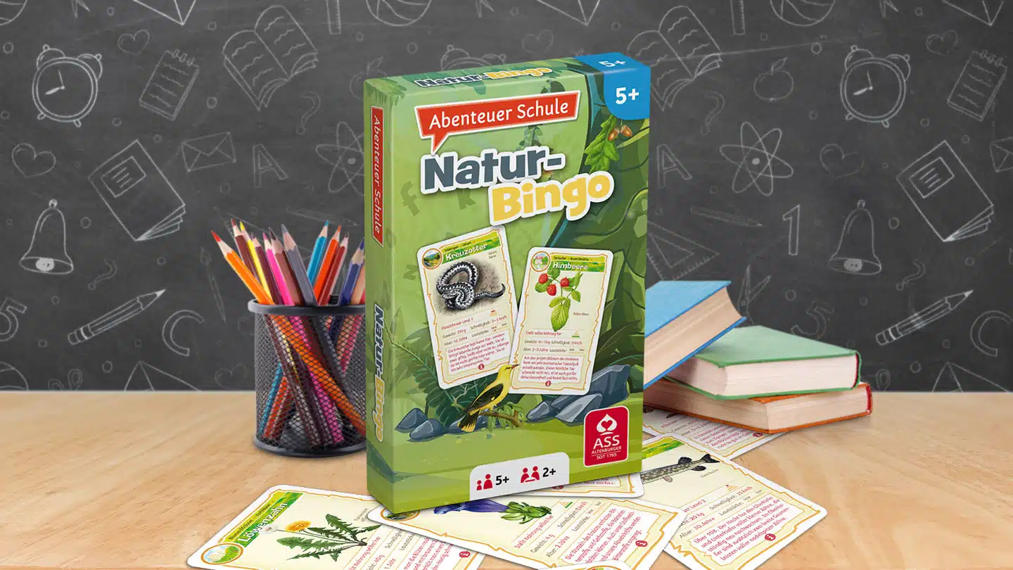 Das Abenteuer Schule Naturbingo Spiel steht auf einem Schultisch. Unter dem Tisch liegen einige der dazugehörigen Spielkarten. Neben dem Spiel stehen Stifte und ein Bücherstapel. Im Hintergrund ist unscharf eine Tafel zu erkennen.