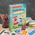 Das Abenteuer Schule Verkehrszeichen Spiel steht auf einem Schultisch. Unter dem Tisch liegen einige der dazugehörigen Spielkarten. Neben dem Spiel stehen Stifte und ein Bücherstapel. Im Hintergrund ist unscharf eine Tafel zu erkennen.