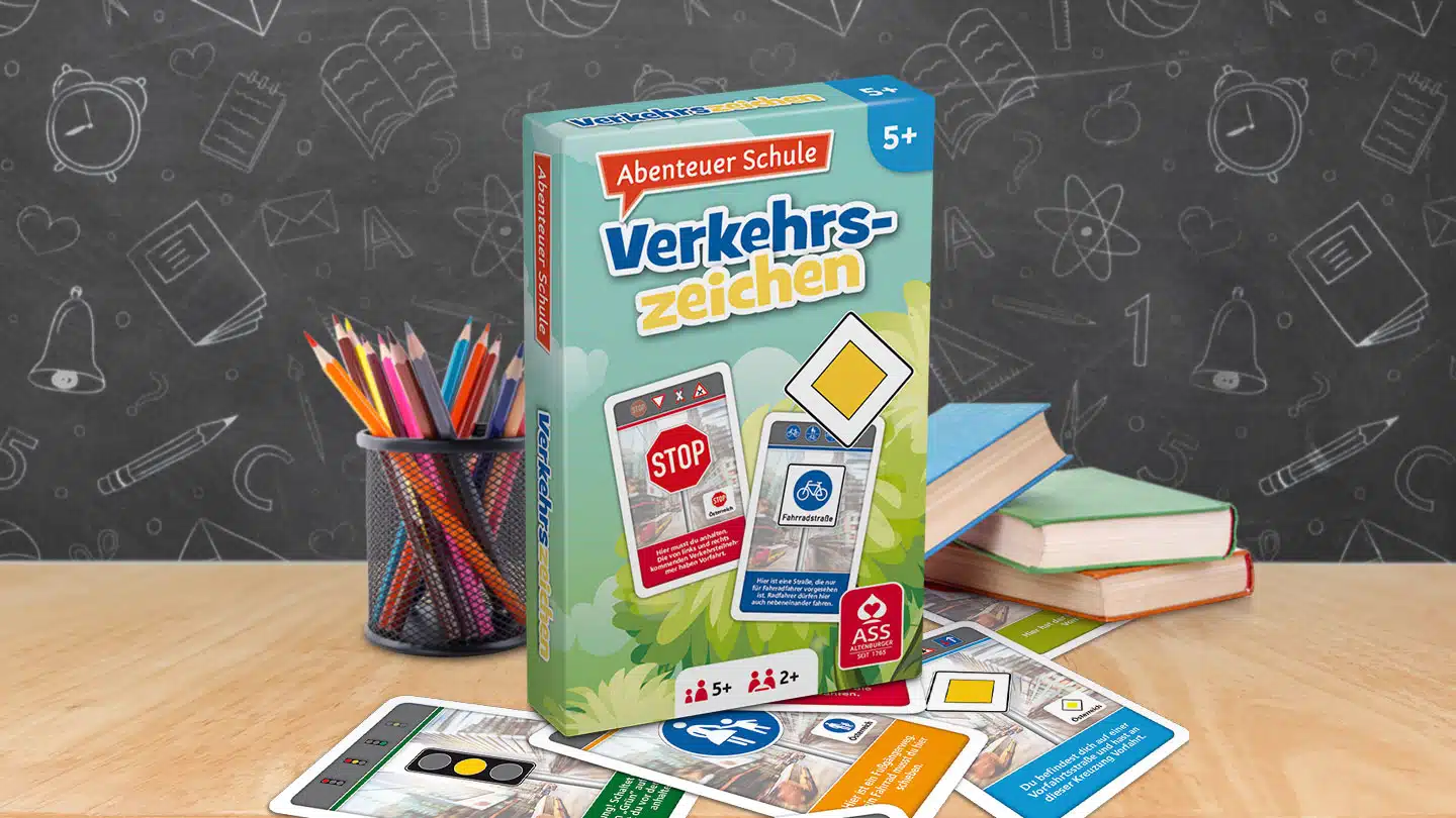 Das Abenteuer Schule Verkehrszeichen Spiel steht auf einem Schultisch. Unter dem Tisch liegen einige der dazugehörigen Spielkarten. Neben dem Spiel stehen Stifte und ein Bücherstapel. Im Hintergrund ist unscharf eine Tafel zu erkennen.