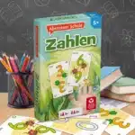 Das Abenteuer Schule Zahlen Spiel steht auf einem Schultisch. Unter dem Tisch liegen einige der dazugehörigen Spielkarten. Neben dem Spiel stehen Stifte und ein Bücherstapel. Im Hintergrund ist unscharf eine Tafel zu erkennen.