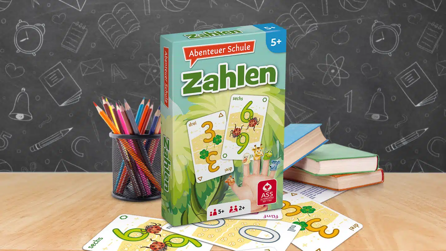 Das Abenteuer Schule Zahlen Spiel steht auf einem Schultisch. Unter dem Tisch liegen einige der dazugehörigen Spielkarten. Neben dem Spiel stehen Stifte und ein Bücherstapel. Im Hintergrund ist unscharf eine Tafel zu erkennen.