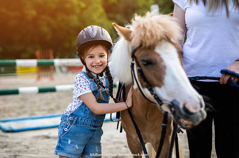 Mädchen mit Reiterhelm steht neben Pony und lächelt. Urlaub auf dem Reiterhof, ASS Altenburger