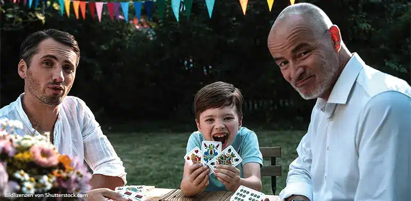 Ein älterer Mann, ein jüngerer Mann und ein Junge sitzen am Gartentisch und spielen Karten. Kartenspiel Knack, ASS Altenburger