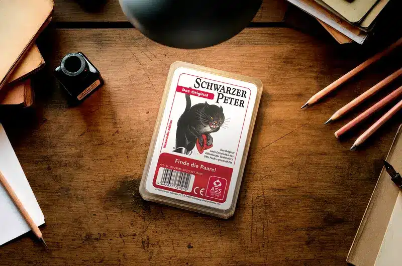 Kartenspiel "Schwarzer Peter" mit schwarzer Katze auf Holztisch umgeben von Stiften, Tintenfass und Notizbüchern. Schwarzer Peter, ASS Altenburger