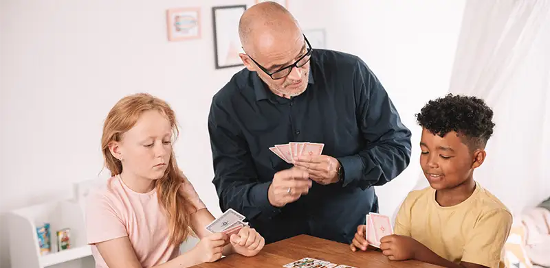 Ein Mann und zwei Kinder sitzen am Tisch und spielen Karten. Bube, Dame, König, ASS Altenburger